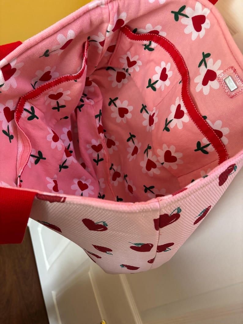 May include: Pink tote bag with a red and white cherry print and a red and white floral print lining. The bag has a red zippered pocket on the inside.