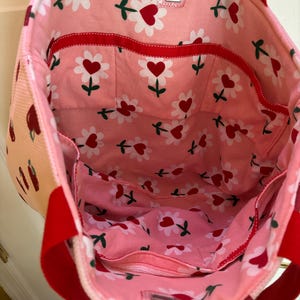 May include: Pink tote bag with red handles and a red trim. The bag has a floral print with white flowers and red hearts. The bag has multiple pockets on the inside.