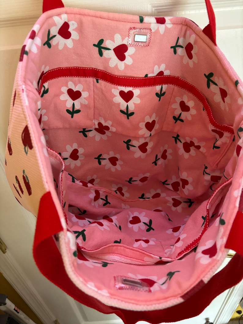 May include: Pink tote bag with red handles and a red trim. The bag has a floral print with white flowers and red hearts. The bag has multiple pockets on the inside.
