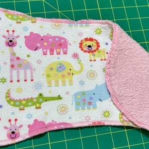 Puede incluir: Un babero para bebé rosa y blanco con un respaldo de felpa suave y absorbente. La parte delantera del babero presenta un estampado colorido de animales, incluidos elefantes, jirafas, hipopótamos y leones.