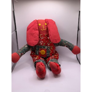 May include: A handmade plush bunny doll with a red head and ears, and a floral print jumpsuit. The bunny has embroidered facial features and a yellow button on the front. The arms and legs are green with a floral pattern and red accents.