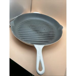 May include: A round white enameled cast iron grill pan with a dark gray interior featuring raised ridges for searing. The handle is white and features the embossed text LE CREUSET. The pan includes two side pouring spouts for liquid drainage.