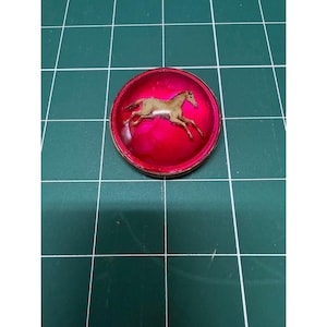 May include: A round, red pin with a brown horse galloping across its surface. The pin has a metallic border and is set against a green background.