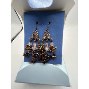 May include: Ornate copper-toned earrings with a vintage aesthetic. Each earring features decorative filigree, small birds, heart-shaped charms, and key-shaped pendants. The earrings also include clear and colorful bead accents, creating a layered and detailed design.