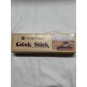 May include: A rectangular box for a Crock Stick knife and scissor sharpener. The box is tan with the words "Crock Stick" in a large, dark brown font. The box also has the words "Knife and Scissor Sharpener".