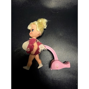 Könnte beinhalten: Eine Vintage-Puppe mit blonden Haaren im Dutt, trägt ein rotes Kleid mit weißer Spitzenborte und einem weißen Blumenmuster. Die Puppe ist an einer rosa Kunststoffbasis befestigt.