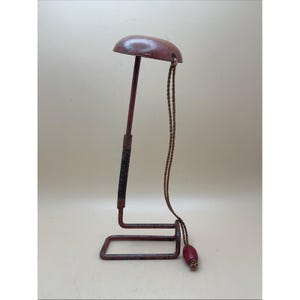 Puede incluir: Un perchero de sombrero vintage con una parte superior semicircular de color granate y una base a juego. El soporte presenta una varilla vertical delgada con una sección negra envuelta y un cordón decorativo con una cuenta granate.