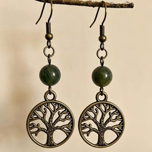 Può includere: Un paio di orecchini pendenti con una perla di pietra rotonda verde scuro sospesa sopra un ciondolo circolare color bronzo che raffigura la silhouette dettagliata di un albero. Gli orecchini sono dotati di ganci in metallo color bronzo.