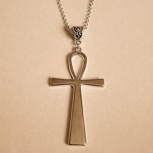 May include: A silver-toned ankh pendant necklace featuring a smooth, polished finish on the cross-shaped charm. The pendant is suspended from a delicate silver-toned chain that connects to the top loop of the ankh with an ornate, filigree-style bail.
