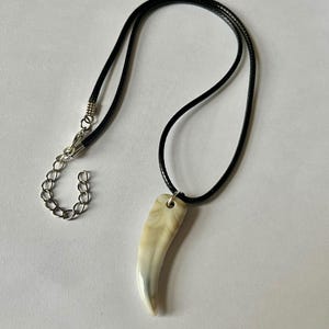 Puede incluir: Un collar con un colgante curvo en forma de diente con un acabado liso jaspeado en crema y bronceado. El colgante cuelga de un cordón negro que incluye un cierre de mosquetón de metal plateado y una cadena de extensión ajustable.