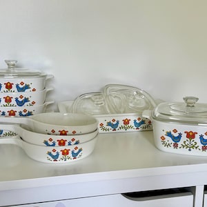 May include: A collection of vintage white ceramic cookware with a blue bird and floral design. The set includes casserole dishes with glass lids, handled bowls, and a rectangular baking dish. The colorful pattern features red, yellow, and green accents.