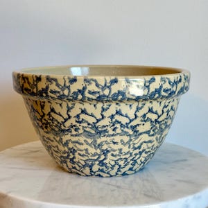 May include: A round ceramic mixing bowl featuring a cream-colored base with a dense, sponge-painted blue pattern covering the exterior. The bowl has a wide, thick rim and a smooth, glossy finish, showcasing a consistent speckled texture throughout.