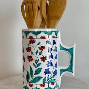 May include: A white ceramic utensil holder with a floral design in red, blue, and green. The holder contains several wooden cooking utensils. The handle and rim are outlined in teal. A decorative kitchen item.