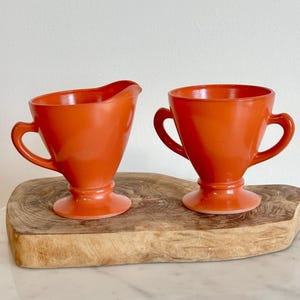 May include: A set of two orange glass creamer and sugar bowls with handles. The bowls have a vintage look and are sitting on a wooden cutting board.