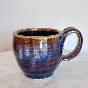 May include: A ceramic mug featuring a rounded body and a curved handle. The exterior and interior display a reactive glaze with deep shades of purple, blue, and brown, creating a layered, variegated appearance with visible horizontal texture.