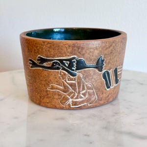 May include: A cylindrical ceramic bowl with a textured, mottled brown exterior featuring an incised, black-and-white graphic illustration of a figure carrying a large bird. The interior of the bowl is finished in a deep, glossy dark green glaze.