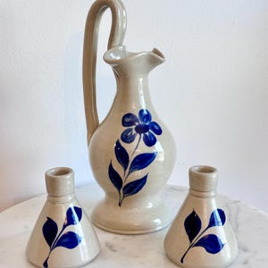 May include: A ceramic pitcher with a tall handle and a ruffled spout, featuring a hand-painted blue flower and leaf design. Two matching conical candle holders with blue leaf motifs are positioned on either side of the pitcher.