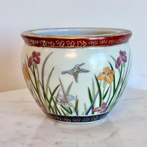 May include: A round ceramic planter with a cream-colored base featuring a hand-painted scene of a small blue bird in flight surrounded by colorful purple, orange, and light blue iris flowers and green grass. The rim is dark red with gold accents.