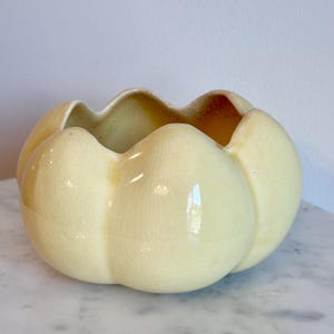 May include: A pale yellow ceramic bowl with a scalloped, flower-like rim and a glossy, crackled glaze finish. The bowl features a rounded, bulbous shape with deep vertical indentations that create a segmented, petal-like appearance throughout.