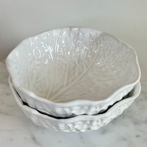 May include: Two white ceramic bowls stacked on top of each other. The bowls are shaped like cabbage leaves, with detailed veining and a textured surface. The bowls are on a white marble surface.