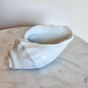 May include: A white ceramic bowl shaped like a seashell with a glossy finish. The vessel features a wide, open top and a textured, ridged exterior that mimics the natural form of a conch shell, with a smooth, curved rim and a hollow interior.