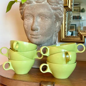 May include: A collection of six lime green teacups with rounded handles, stacked on a wooden surface. The teacups are arranged in two stacks of three. A stone bust of a woman and a gold-framed mirror are in the background.