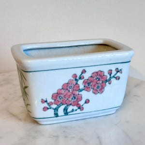 May include: A rectangular white ceramic planter featuring a hand-painted design of pink cherry blossoms on branches with dark green stems. The pot has a smooth, glossy finish and a thin green border line encircling the upper and lower edges.