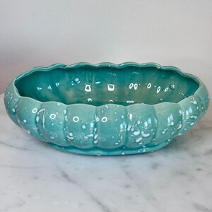 May include: An oval ceramic bowl featuring a scalloped edge and a glossy turquoise glaze with white speckled accents. The bowl has a wide, open interior and a rounded, fluted exterior design, creating a textured appearance across the surface.