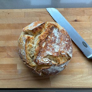 Op de afbeelding: Een rond brood met een goudbruine korst, bestrooid met bloem, ligt op een houten snijplank. Een gekarteld broodmes met een zwart handvat ligt ernaast, klaar om te snijden.