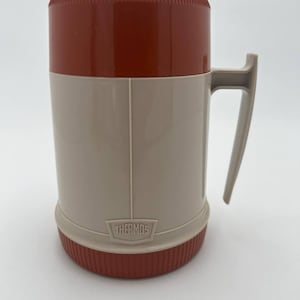May include: A vintage Thermos brand food jar featuring a beige cylindrical body with a matching side handle. The top lid and the bottom base are both a deep reddish-brown colour with a ribbed texture. The word THERMOS is embossed on the front.