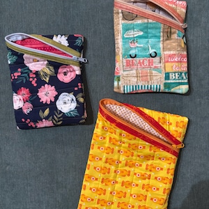 May include: Three zippered pouches with different patterns. The first pouch is navy blue with pink and white flowers. The second pouch is beige with a turquoise background and a scooter, palm trees, and the words "Beach" and "Welcome to the Beach". The third pouch is yellow with a pattern of orange fish.