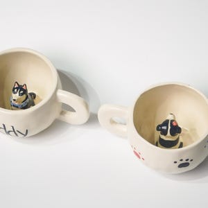 Könnte beinhalten: Zwei cremefarbene Keramikbecher mit Hundefiguren im Inneren. Auf einem Becher steht das Wort "puppy". Der andere Becher hat Pfotenabdrücke und eine Hundefigur. Beide Becher haben Henkel.