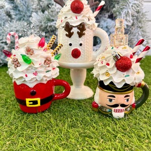 May include: Three decorative mugs topped with faux whipped cream. One is a red Santa suit mug, one is a white reindeer mug on a pedestal, and one is a nutcracker mug. Each features festive embellishments like candy canes, ornaments, and lights.
