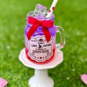 May include: A mason jar filled with a purple and red liquid, ice cubes, and a straw with red hearts. The jar has a red bow and a label that reads "Cupid's Love Potion." It sits on a white pedestal on a green surface.