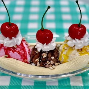 May include: A classic banana split dessert in a clear glass dish. It features scoops of pink, brown, and yellow ice cream, topped with whipped cream and a bright red cherry. The split sits on a halved banana.