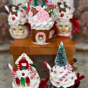 May include: A collection of Christmas-themed mugs with whipped cream toppings and festive decorations. The mugs feature Santa faces, gingerbread men, candy canes, and miniature Christmas trees. The color palette includes red, white, brown, and green.