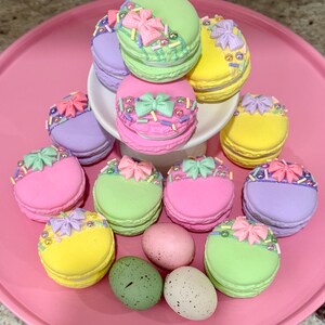 May include: A pink plate with a variety of pastel-colored macarons decorated with bows and sprinkles. There are also three speckled Easter eggs on the plate.
