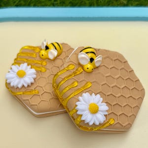 May include: Two hexagonal honeycombs with a yellow and black bee on each. The honeycombs are decorated with yellow honey drips and white daisies.