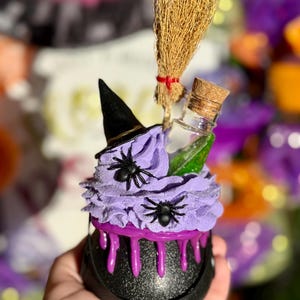 May include: A Halloween-themed decoration featuring a black cauldron with purple frosting and dripping accents. It's topped with a witch's hat, spiders, a miniature broom, and a small bottle. The color scheme includes black, purple, and brown.