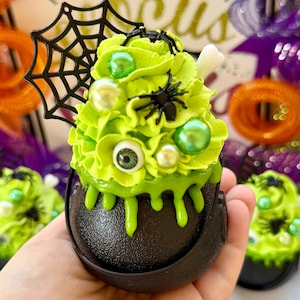 May include: A black cauldron-shaped container filled with lime green frosting, decorated with green beads, a white bone, and black spiders. A black spiderweb is attached to the cauldron. The text in the background says "It's just a bunch of focus."