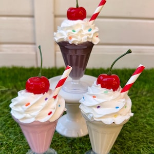May include: Three decorative milkshake glasses with whipped cream, sprinkles, a cherry, and a striped straw. One glass is pink, one is beige, and one is dark brown. The glasses are on a white pedestal and green grass.