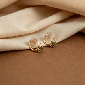 May include: A pair of gold-toned hoop earrings featuring a small, round, faceted olive-green gemstone set in a prong setting. Each earring includes a clear, circular silicone backing attached to the curved metal post for secure placement.