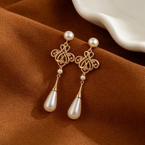 May include: A pair of gold-tone drop earrings featuring a small white pearl stud at the top, connected to an intricate gold filigree scrollwork design with embedded clear stones, and finished with a dangling white teardrop-shaped pearl pendant.