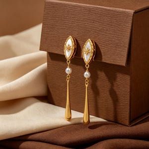 Könnte beinhalten: Ein Paar goldfarbene Ohrhänger mit einem marquiseförmigen Oberteil, einer weißen Perlmutt-Einlage und einem zentralen Sternmotiv. Jeder Ohrring hat eine kleine weiße Perle, die über einem glatten, länglichen, goldfarbenen Tropfenanhänger hängt.