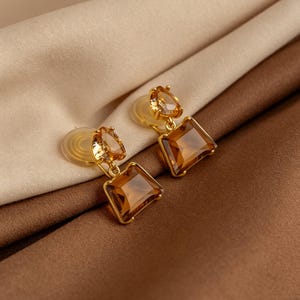 May include: A pair of gold-toned clip-on earrings featuring a small, faceted, amber-colored teardrop stone at the top, connected by a metal link to a larger, trapezoidal, faceted amber-colored stone at the bottom, with clear plastic backings.
