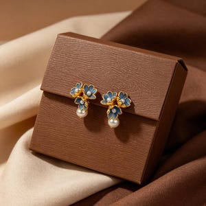 Könnte beinhalten: Ein Paar goldfarbene Ohrstecker in Form von dreiblättrigen blauen Blumen mit goldenen Rändern. Jeder Ohrring hat eine kleine weiße Perle in der Blütenmitte und eine größere weiße Perle, die unter der Blumenform herabhängt.