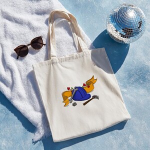 Può includere: Una borsa tote color crema con un casco blu, una chiave inglese, un martello e uno striscione con rose rosse. La borsa è su un asciugamano bianco con occhiali da sole e una palla da discoteca sullo sfondo.