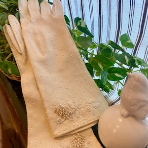 May include: A pair of white long gloves with pearl beading. The gloves are made of a soft, textured fabric and have a delicate floral design.