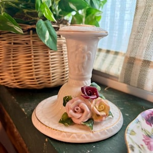 May include: A white porcelain candlestick holder with a decorative floral design. The base features three ceramic roses in pink, yellow, and burgundy, with green leaves. The rim is trimmed with gold. A wicker basket and green plant are in the background.