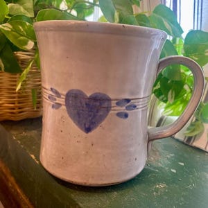 Könnte beinhalten: Eine weiße Keramiktasse mit einem blauen Herzen und blauen Akzenten. Die Tasse hat einen Henkel und steht auf einer grünen Oberfläche.